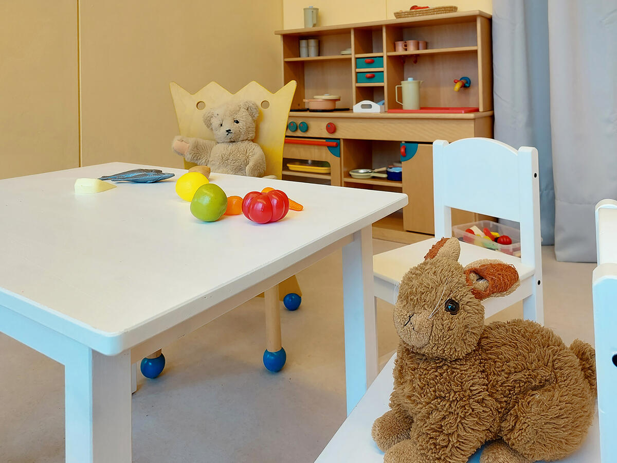 Kuscheltiere sitzen am Tisch. Im Hintergrund ist eine Spielküche aus Holz