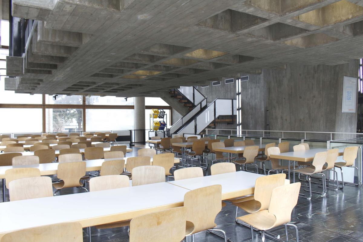 Dining room of the Kunstakademie canteen. Numerous tables and chairs. At the back right is a staircase to the next floor.