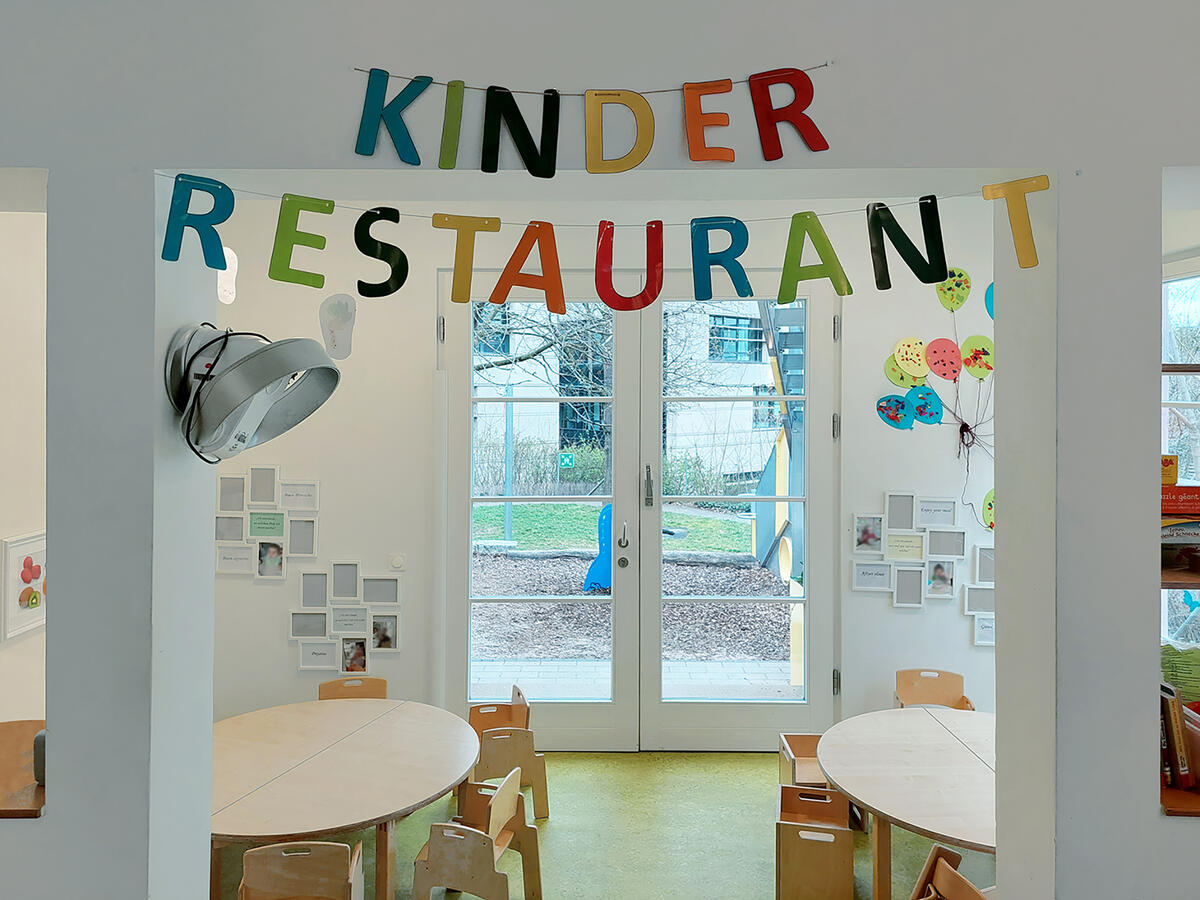 Essbereich der Kita Villa MiO mit Holztischen und -stühlen. Buchstabengirlande mit "Kinderrestaurant"